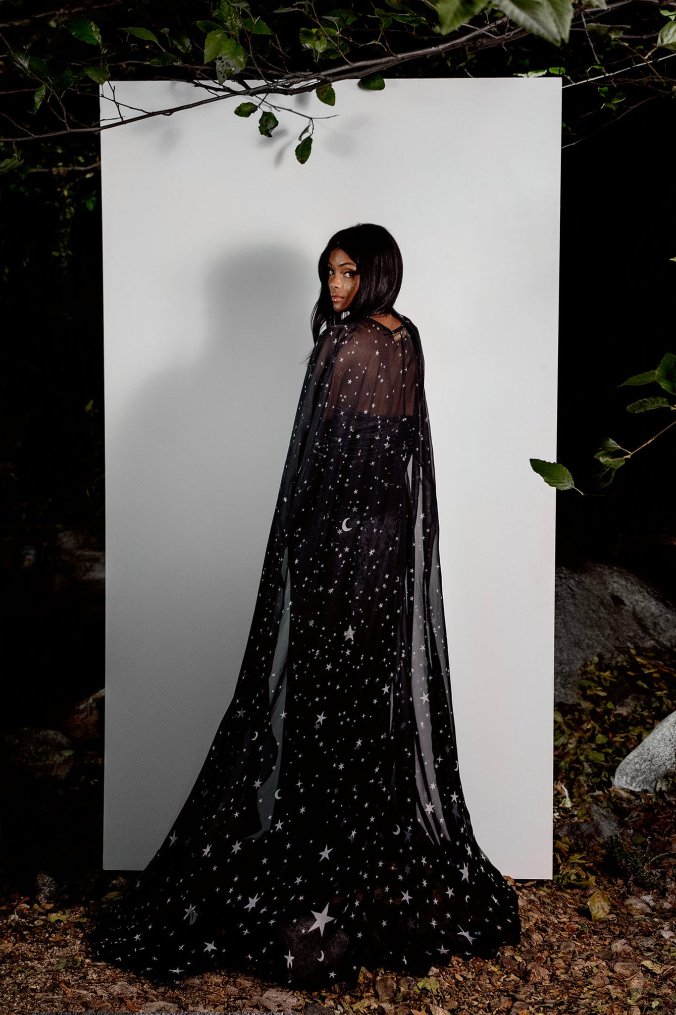 Person wearing a black star-patterned cape against a white background with natural elements around.