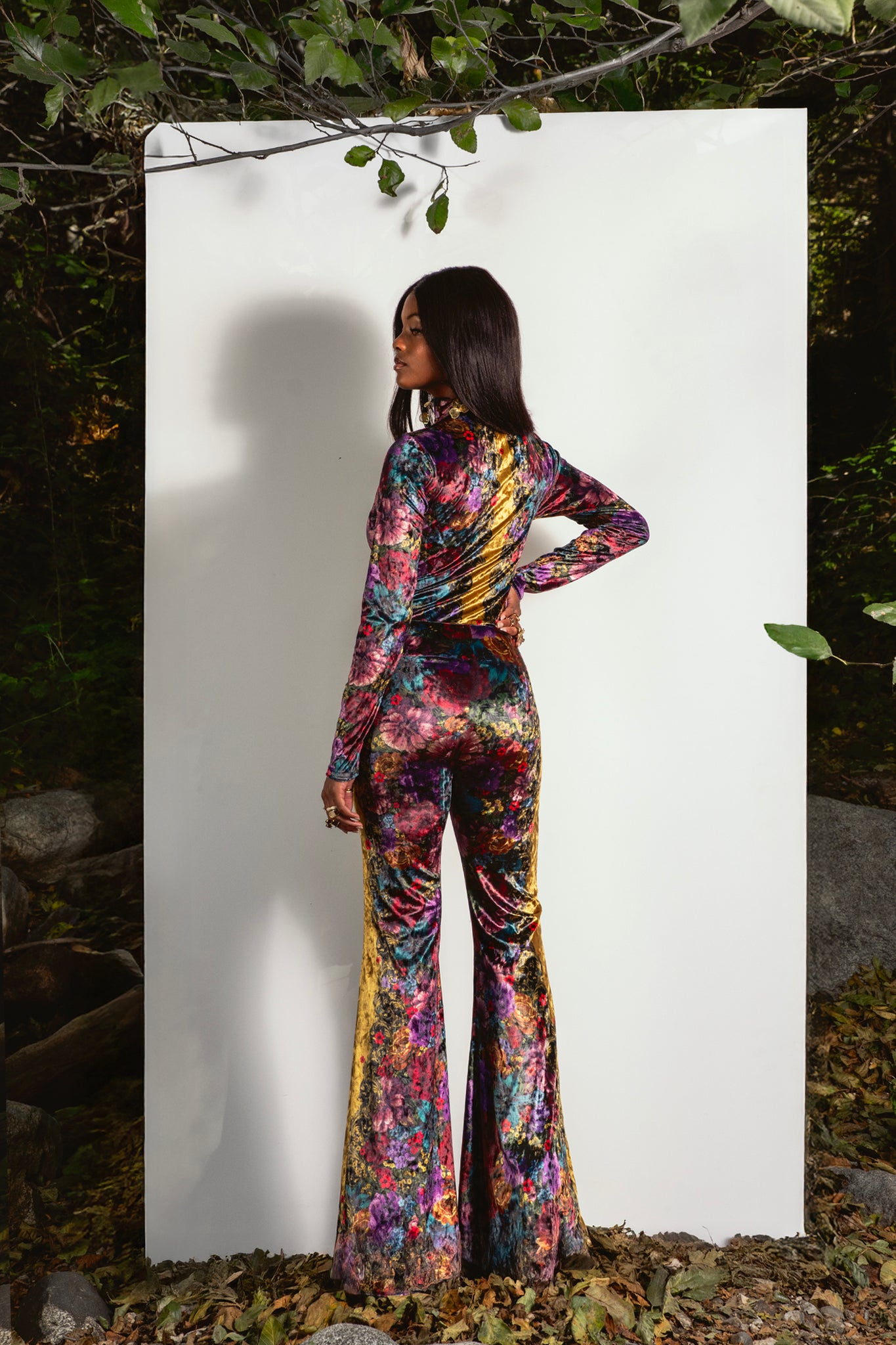 Person wearing a floral bell pant and bodysuit standing against a white background with greenery around.