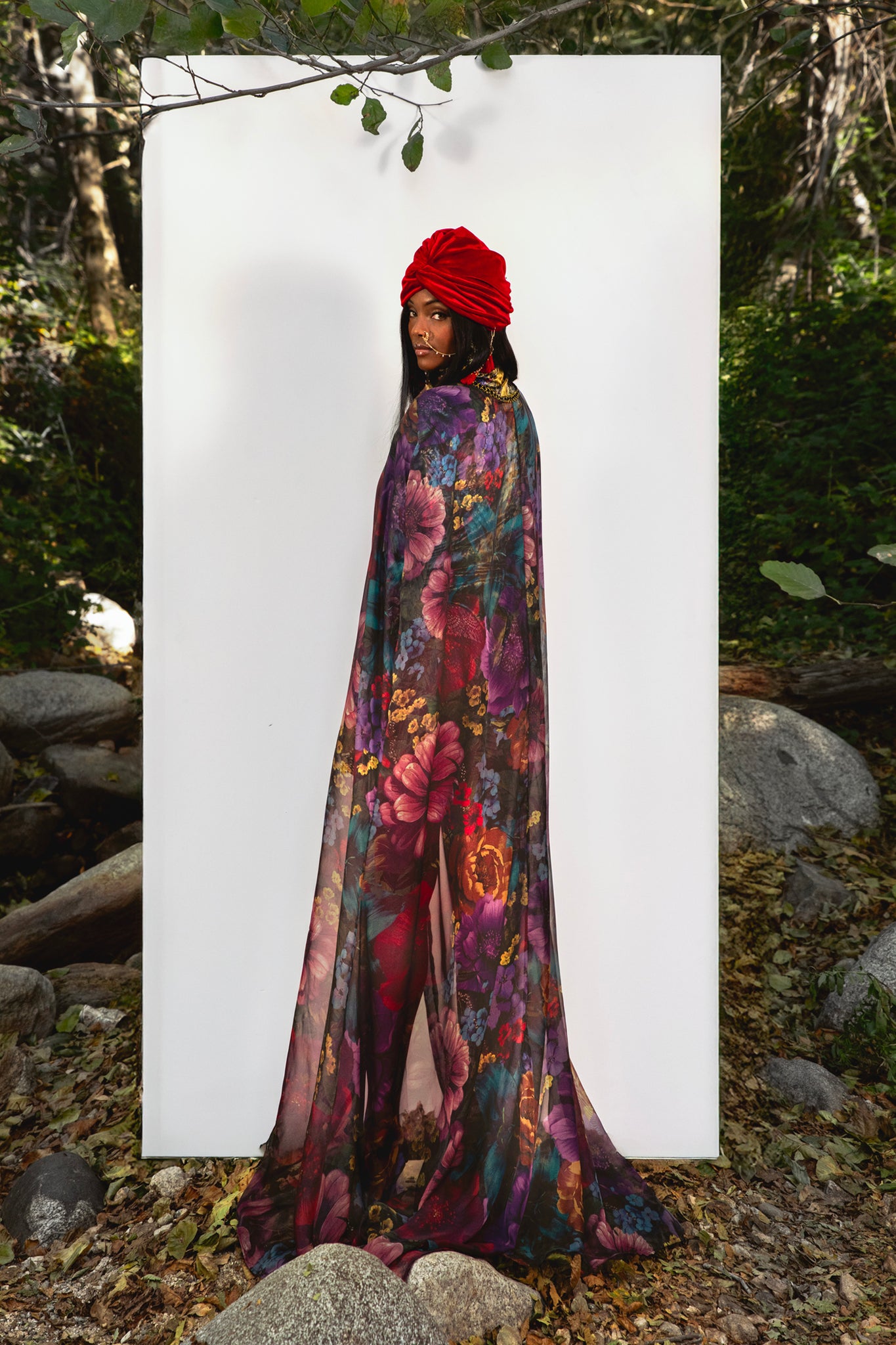Person wearing a colorful floral cape and red headscarf standing against a white background with a natural setting.