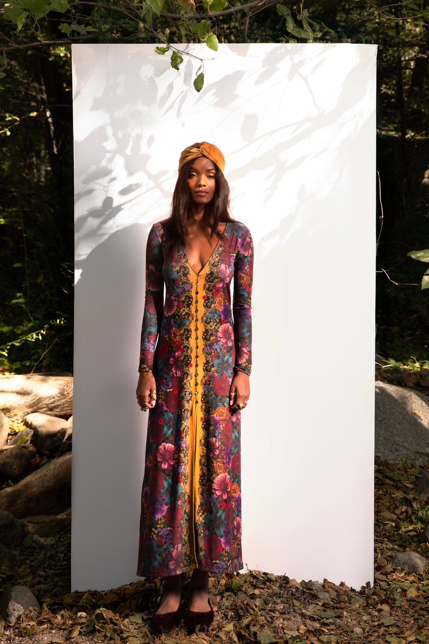 Woman wearing a colorful floral dress and headscarf standing against a white backdrop in a natural setting.