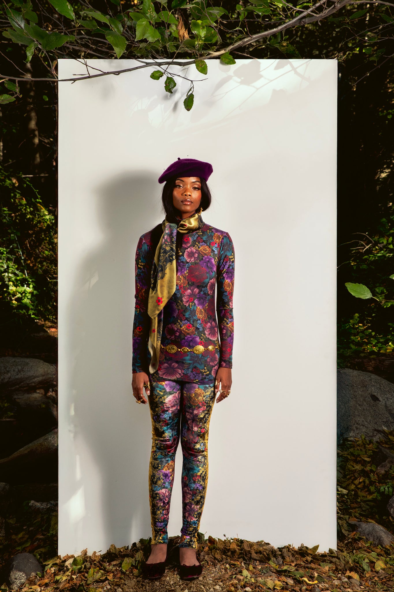 Person wearing a colorful outfit with a floral pattern, standing against a white background with greenery around.