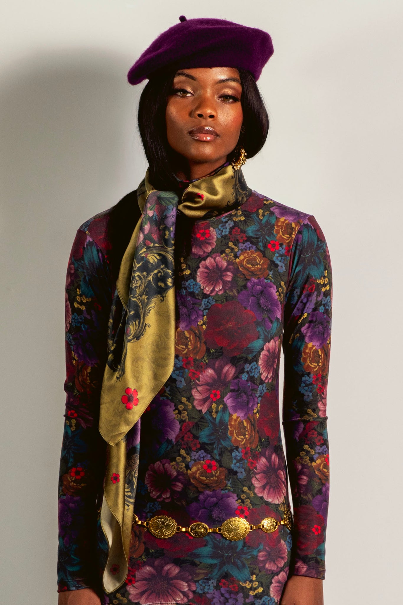Woman wearing a floral dress with a purple beret and gold scarf on a plain background