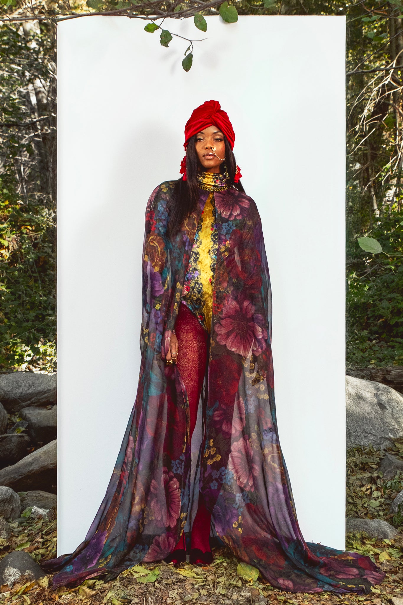 Person wearing a colorful floral cape and red headscarf standing against a white background with a natural setting.