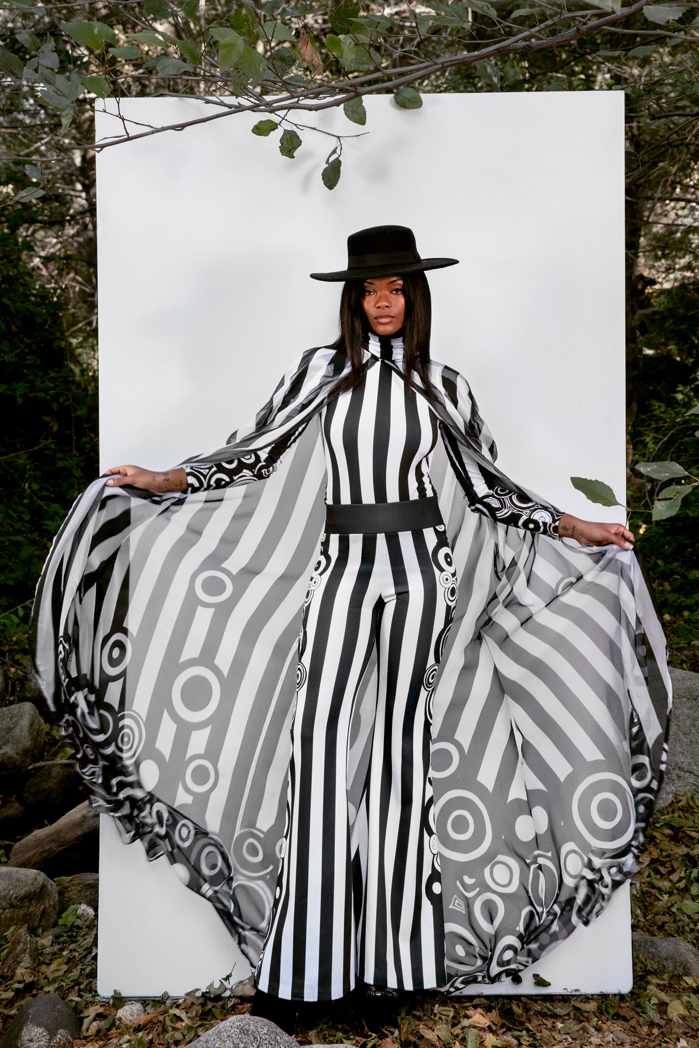 Person wearing a black and white bodysuit with a hat, standing against a white backdrop outdoors.
