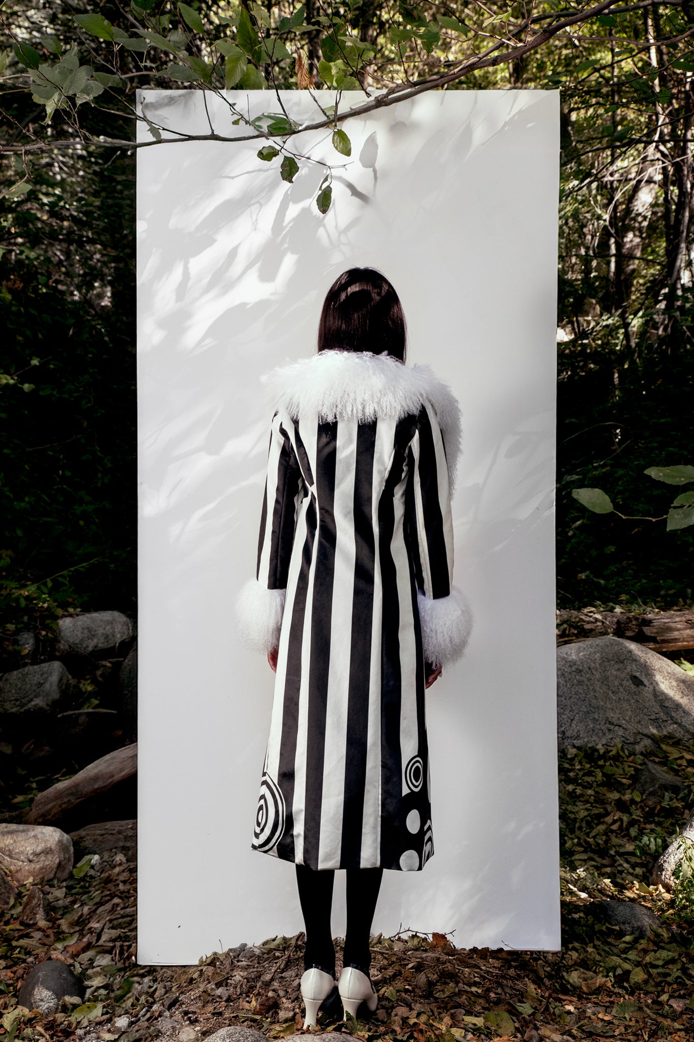 Person wearing a black and white striped coat with fur trim standing against a white board in a forest setting.