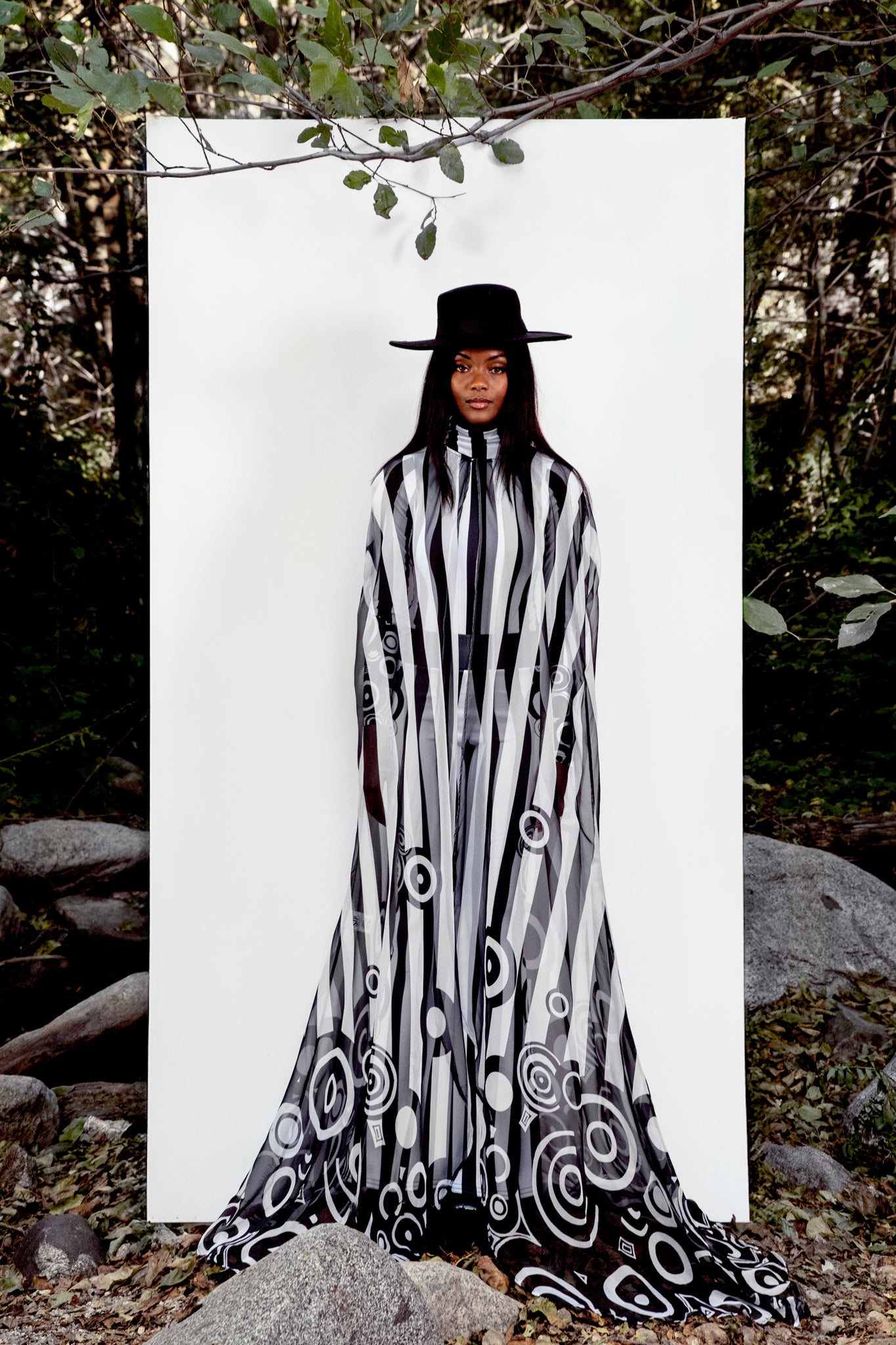 Person wearing a black and white patterned cape with a hat in a forest setting
