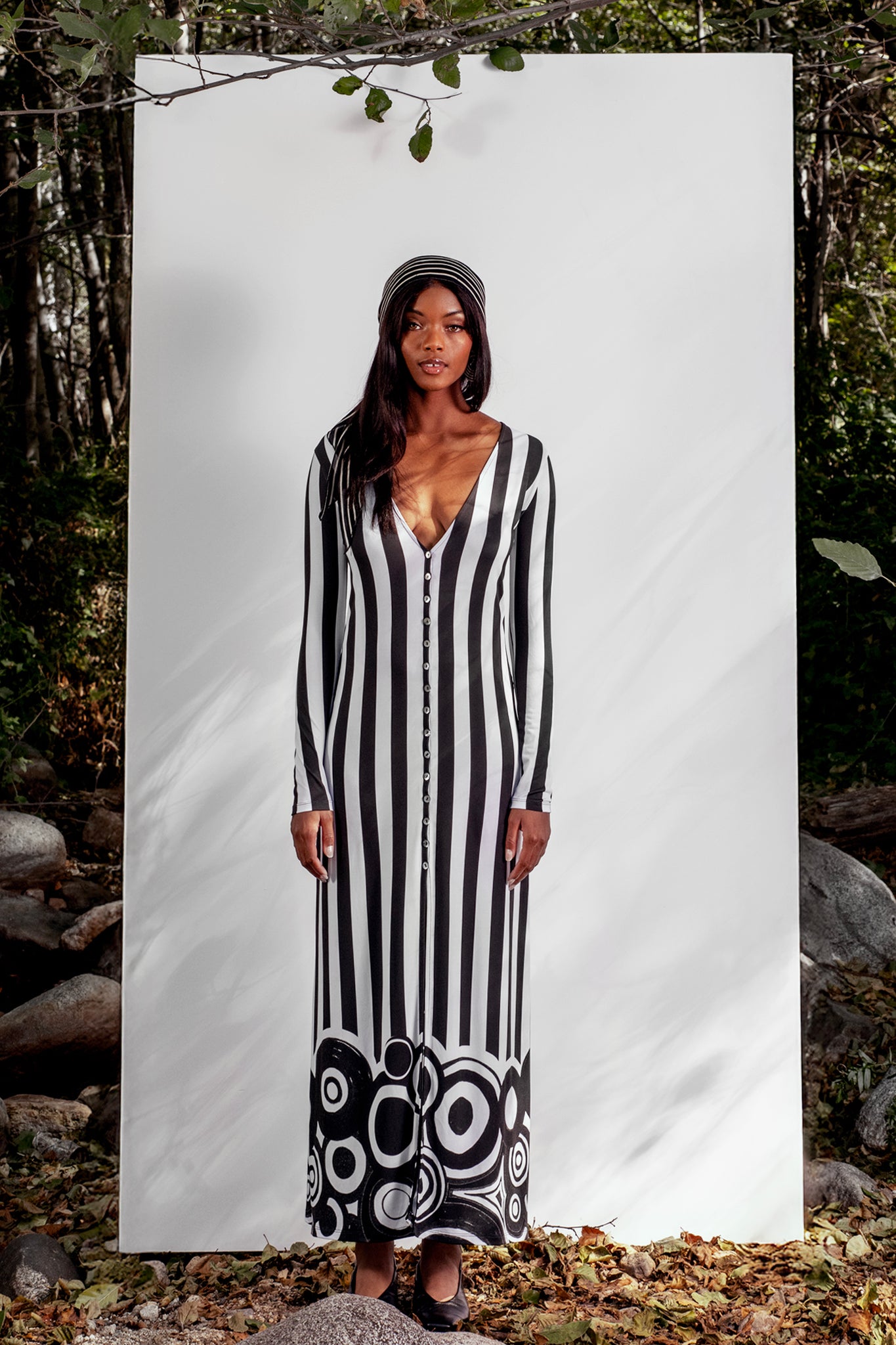 Woman wearing a black and white striped dress with a patterned bottom section, standing against a white background with nature around.