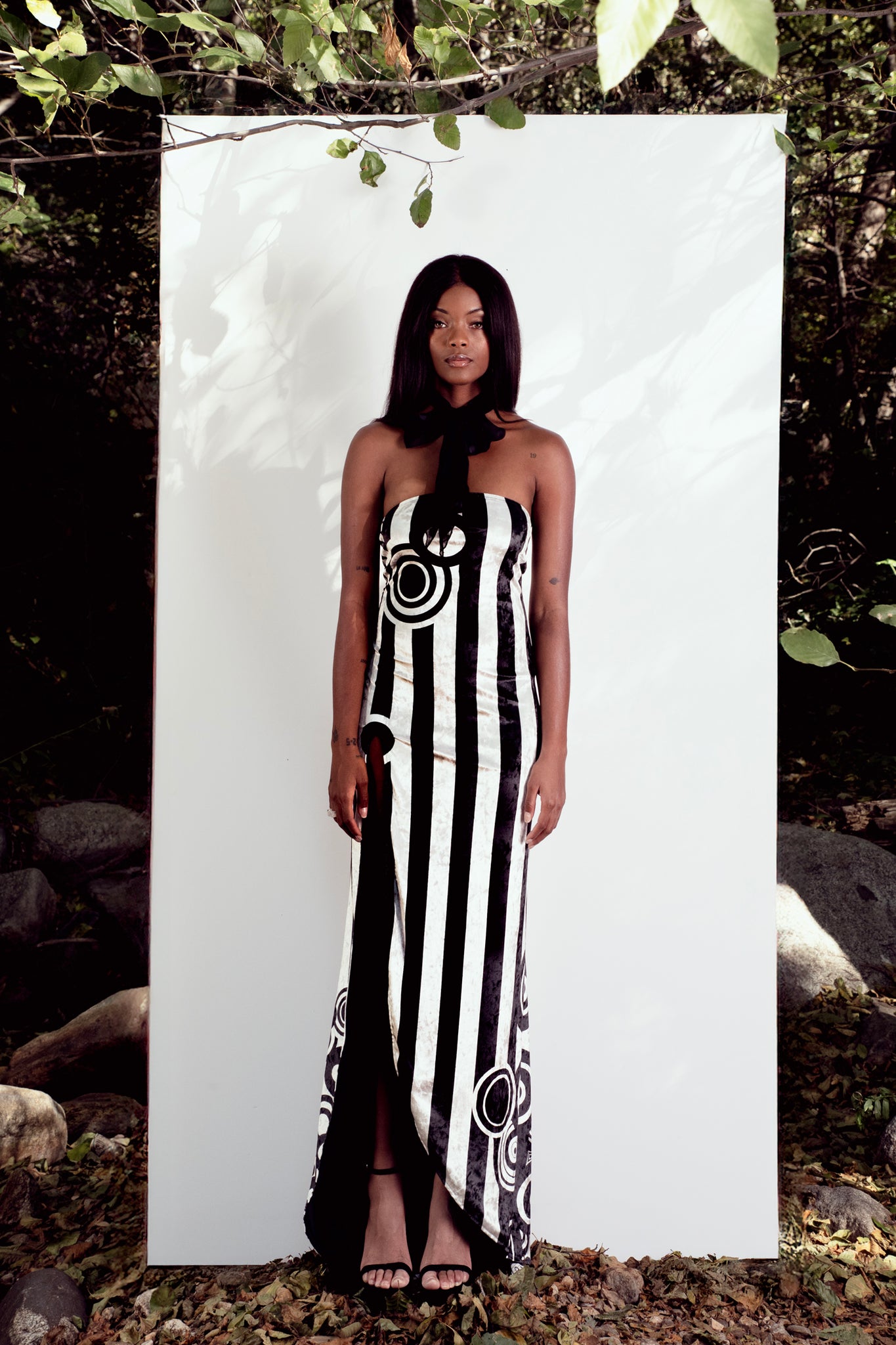 Woman wearing a black and white patterned dress standing against a white backdrop with nature around.